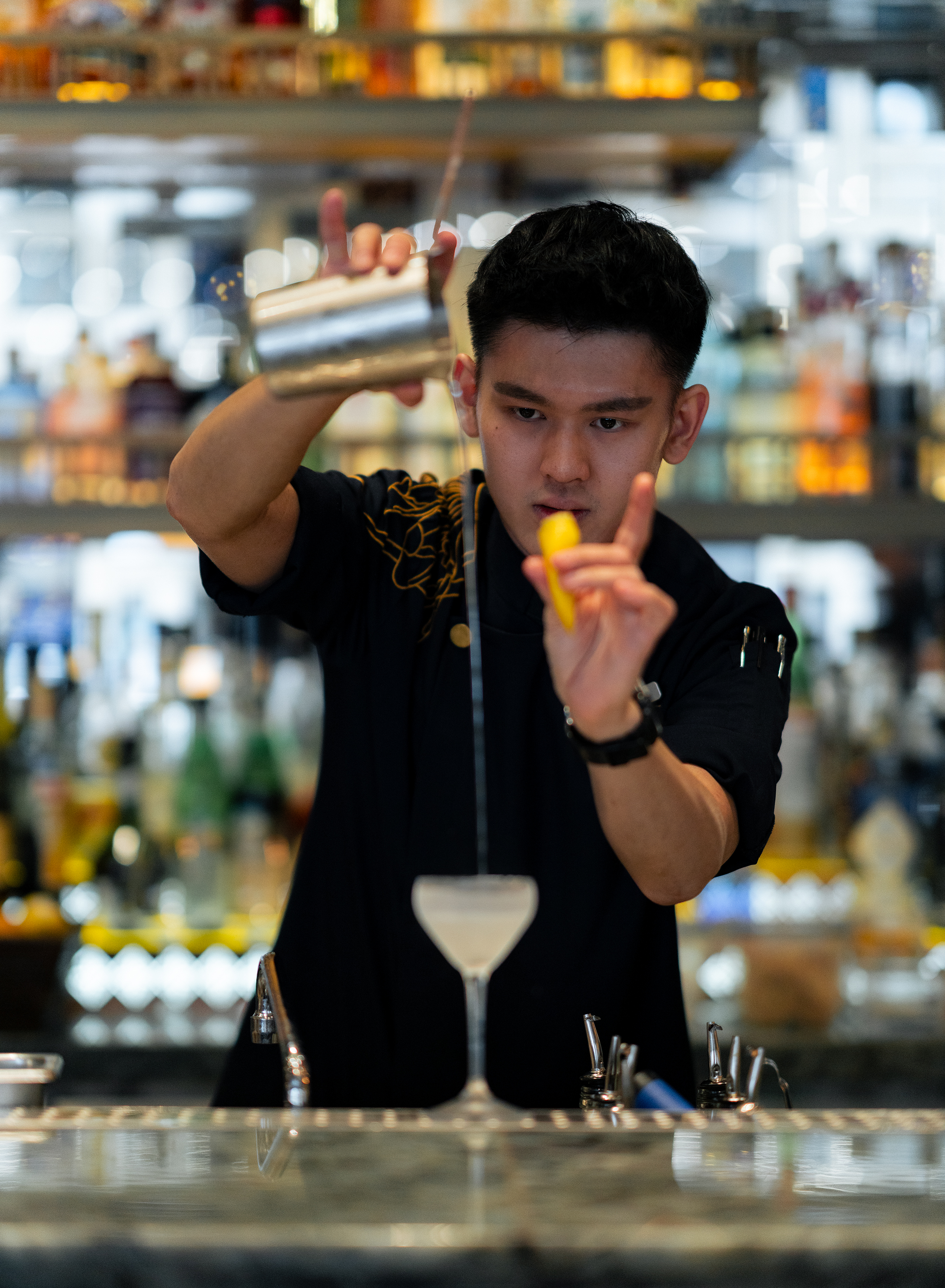 A focused bartender at the Four Seasons Kuala Lumpur expertly pours a pale yellow cocktail through a fine stream into a chilled coupe glass, holding a lemon twist garnish in one hand against a backdrop of blurred shelves stocked with premium spirits