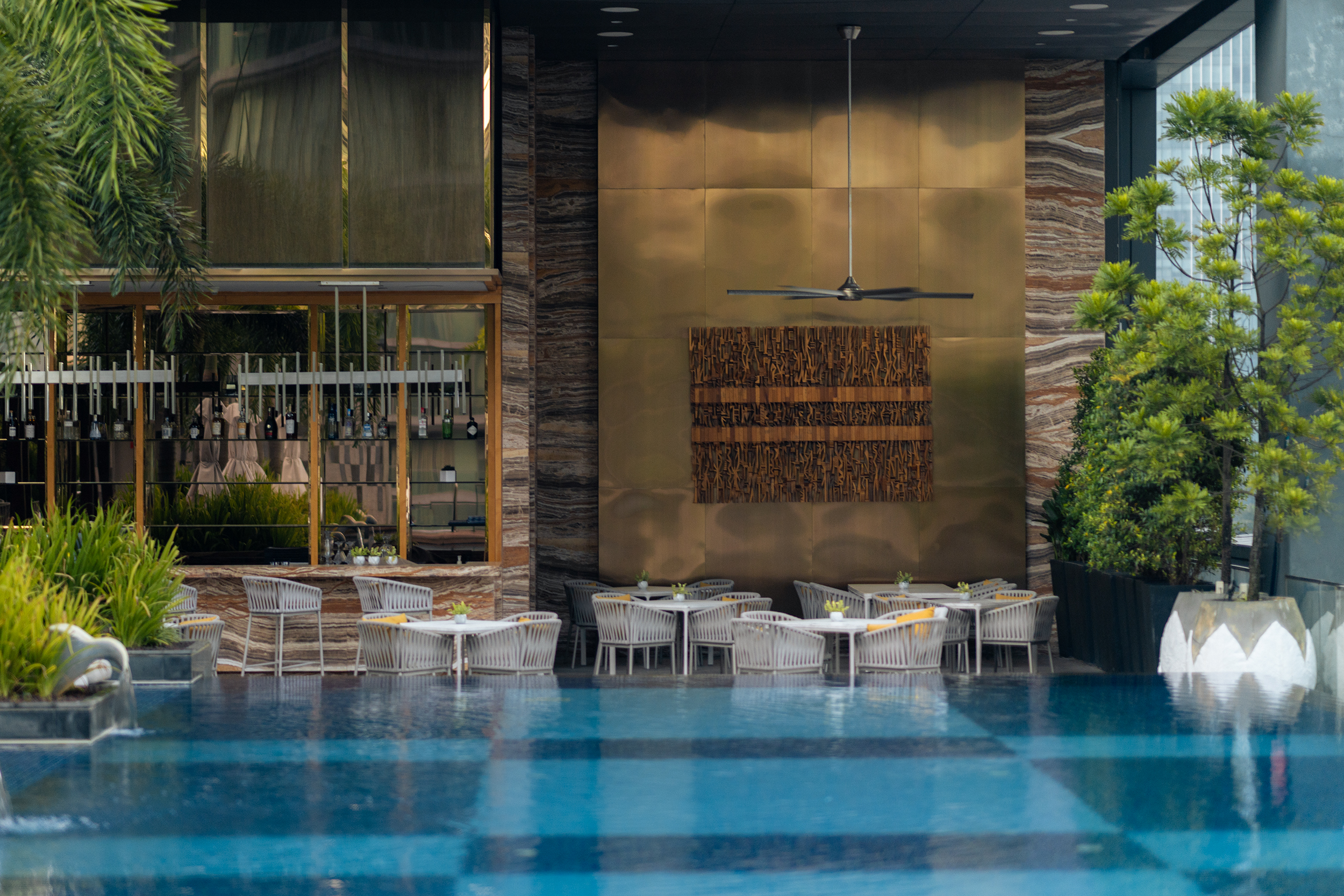 The Four Seasons Kuala Lumpur pool bar with wicker dining chairs arranged around white-topped tables alongside a blue-tiled infinity pool. Golden bronze panels and a sculptural wall art piece form the backdrop, while lush potted greenery frames the scene