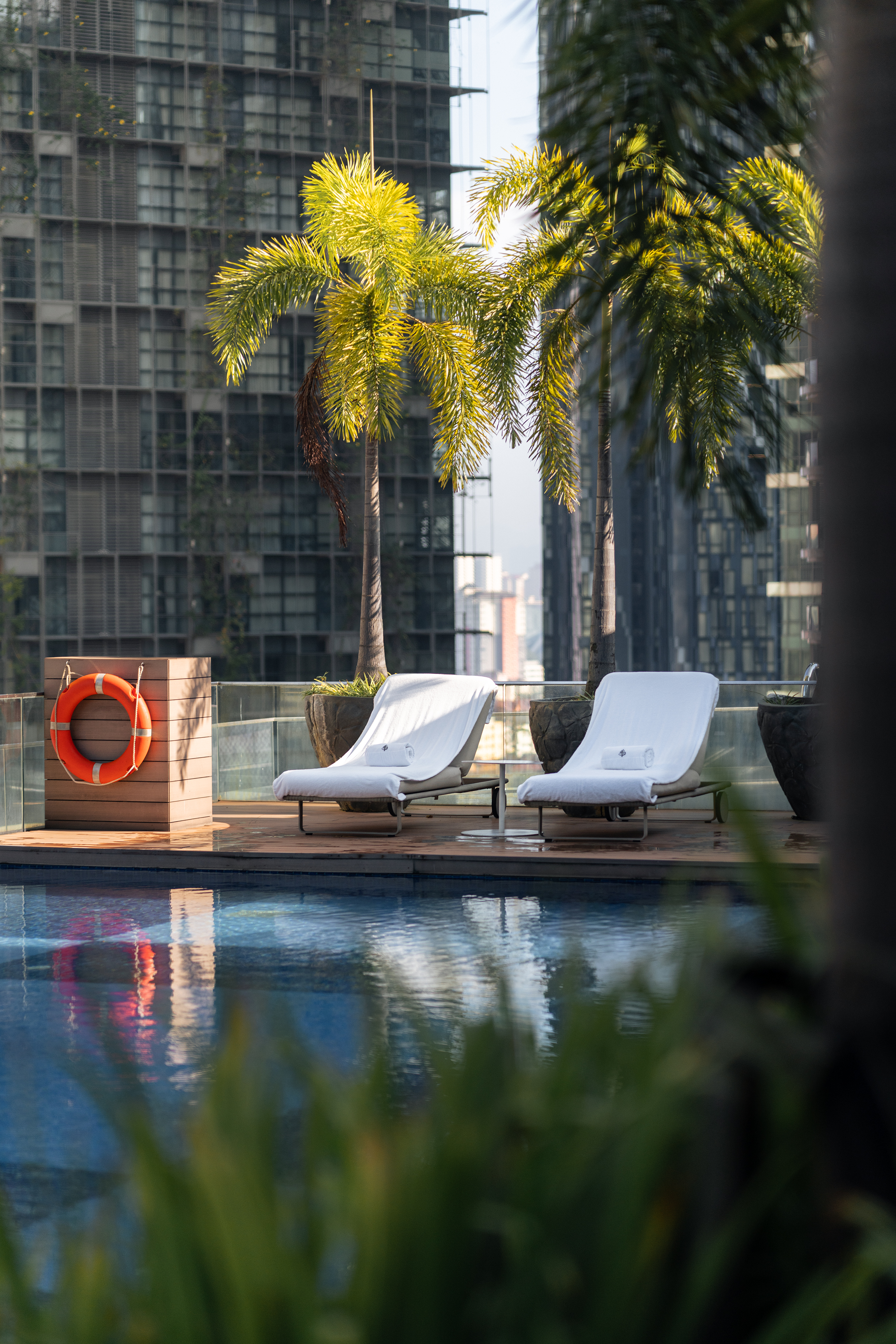 A rooftop swimming pool at the Four Seasons Kuala Lumpur with two white-cushioned sun loungers and a life ring mounted on a wooden planter box. Tall palm trees rise above, and city skyscrapers reflect off the water’s surface.
