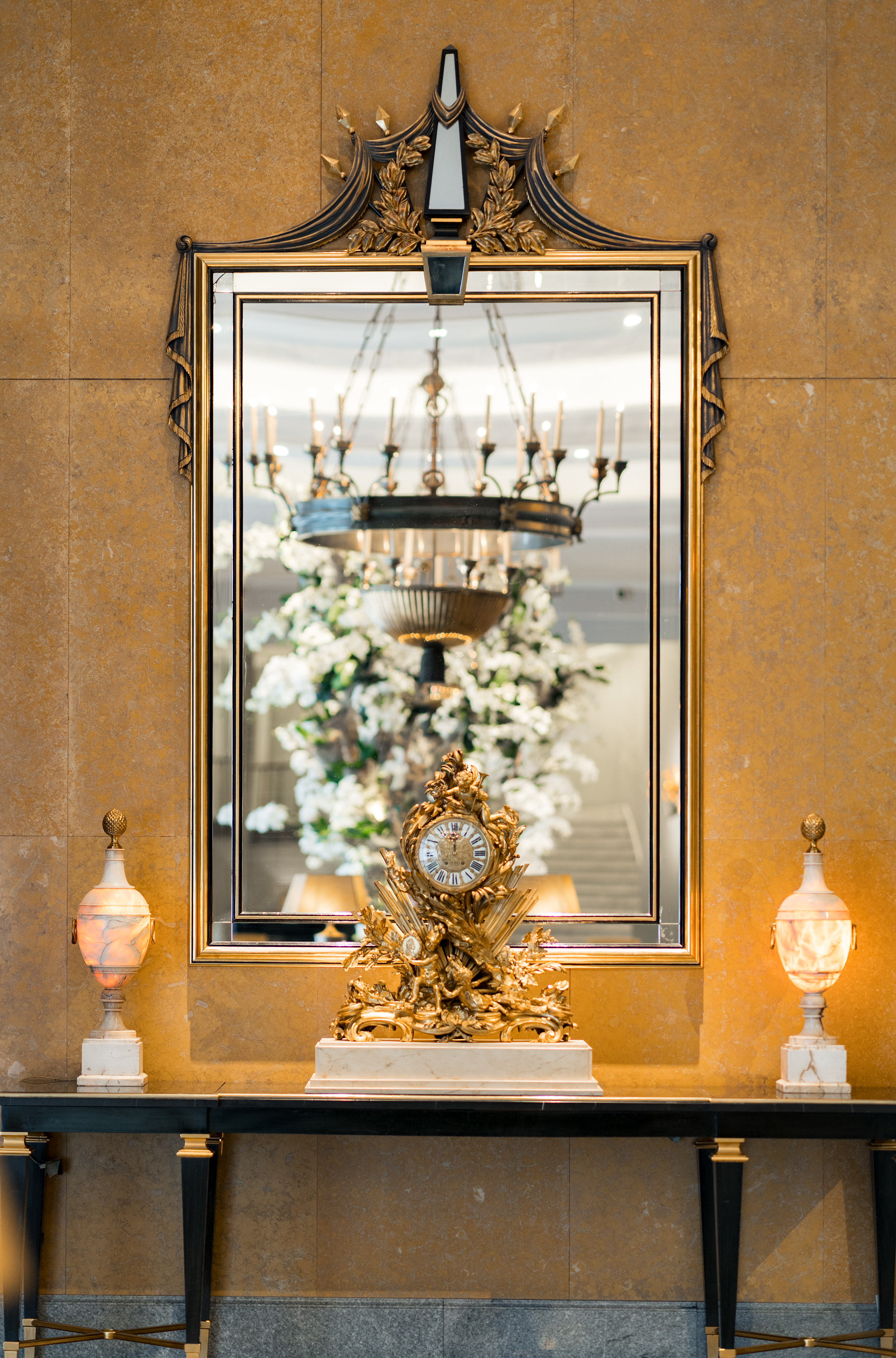 An ornate gilt-framed mirror above a black marble console table in the Four Seasons Lisbon lobby, flanked by two alabaster urn lamps. A decorative gold mantel clock sits at center, reflecting a grand floral arrangement behind it