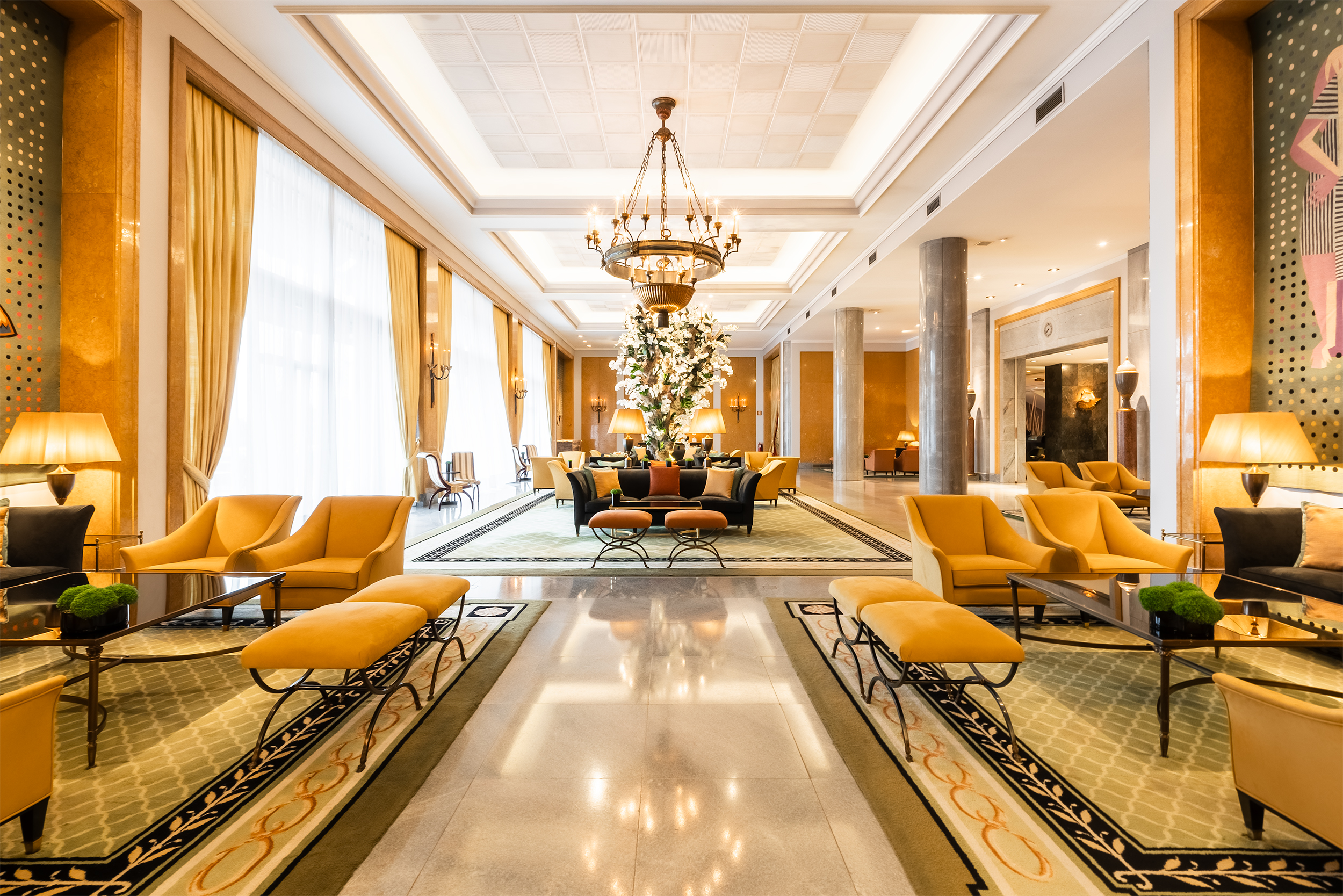 The bright and elegant main lobby of the Four Seasons Lisbon, featuring rows of mustard-yellow lounge chairs and benches on patterned rugs, a central chandelier above a towering white orchid display, and large windows draped with golden curtains that flood the space with natural light