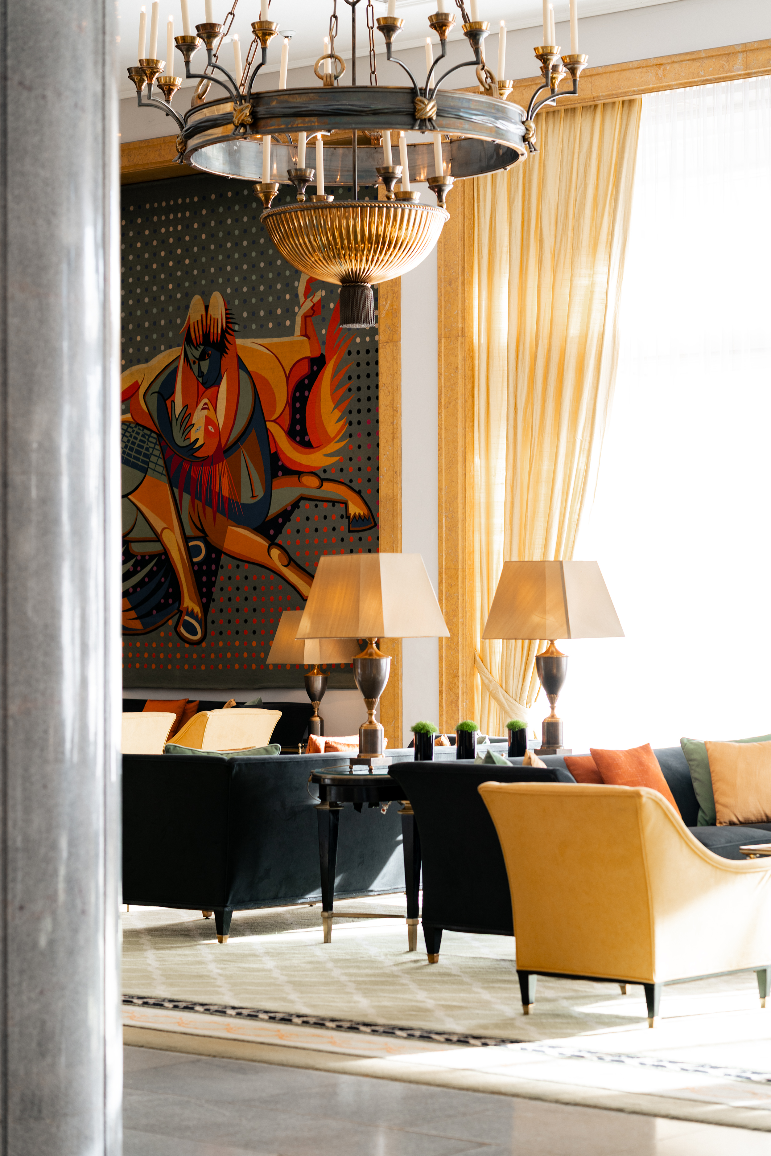 A sunlit hotel lounge at the Four Seasons Lisbon featuring plush velvet sofas in deep green and mustard yellow, accented with brass table lamps and a large modern chandelier above. A vibrant abstract tapestry hangs on the marble-framed wall, while floor-to-ceiling windows drape sheer golden curtains, creating a warm, inviting seating area