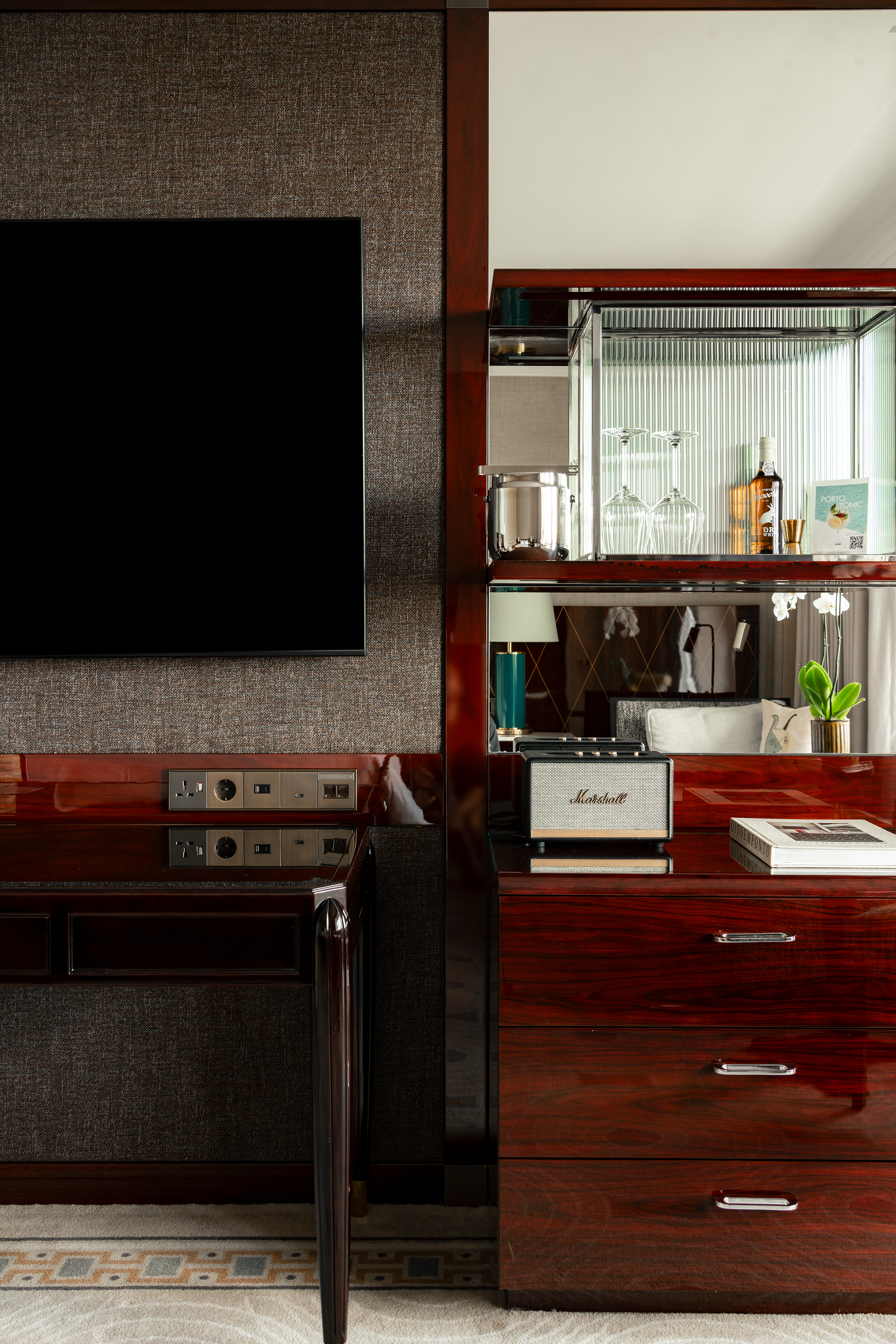 A luxurious Four Seasons Lisbon guest room detail showing a dark wood media console with a mounted flat-screen TV on one side and a mirrored minibar cabinet on the other. Polished surfaces reflect natural light, while a sleek Marshall Bluetooth speaker and neatly arranged glassware hint at upscale in-room amenities