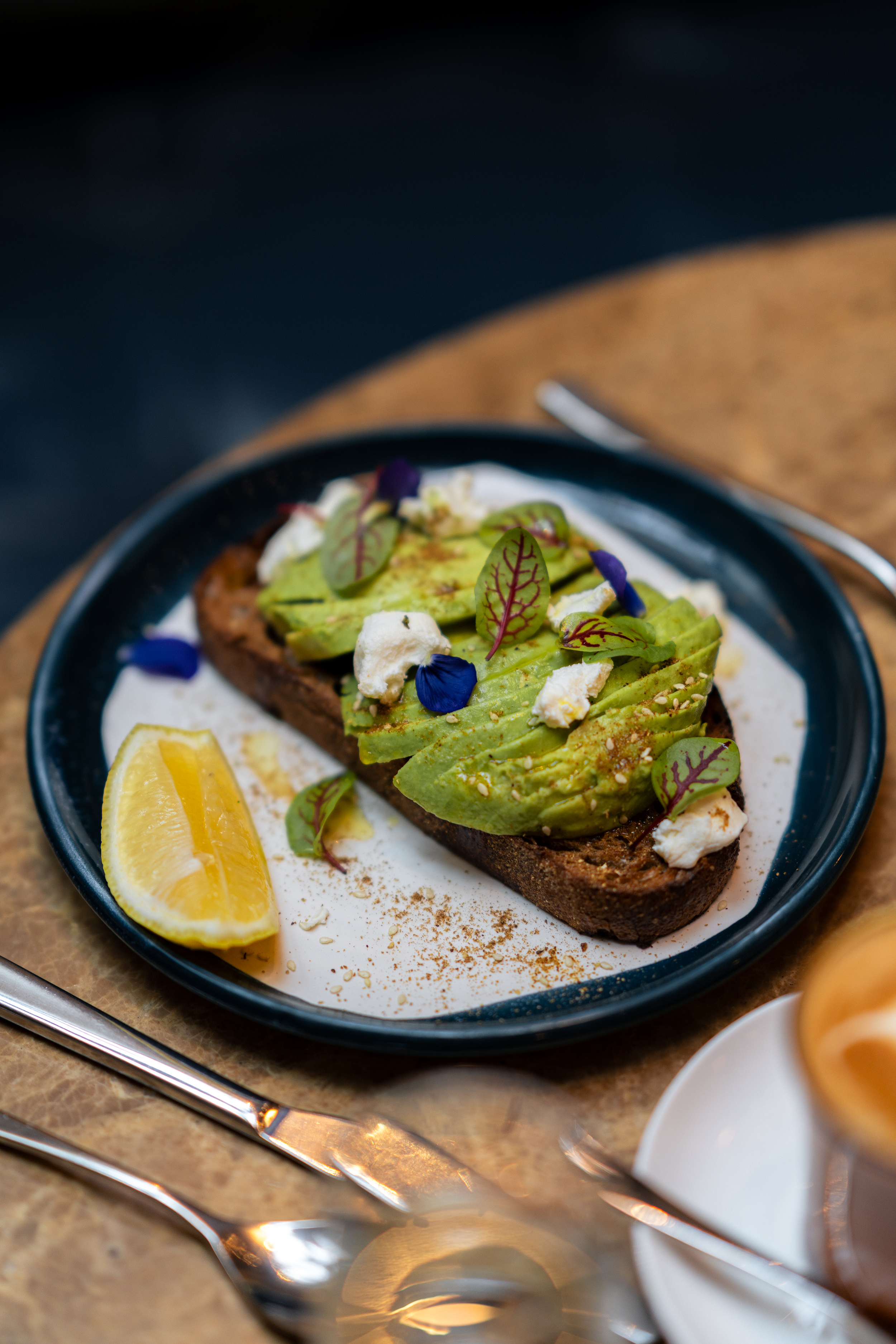 A close-up of a gourmet breakfast serving at the Four Seasons Sydney: a slice of artisan whole-grain toast topped with fanned avocado slices, dollops of whipped ricotta, microgreens, edible petals, and a lemon wedge, presented on a dark ceramic plate alongside polished silverware