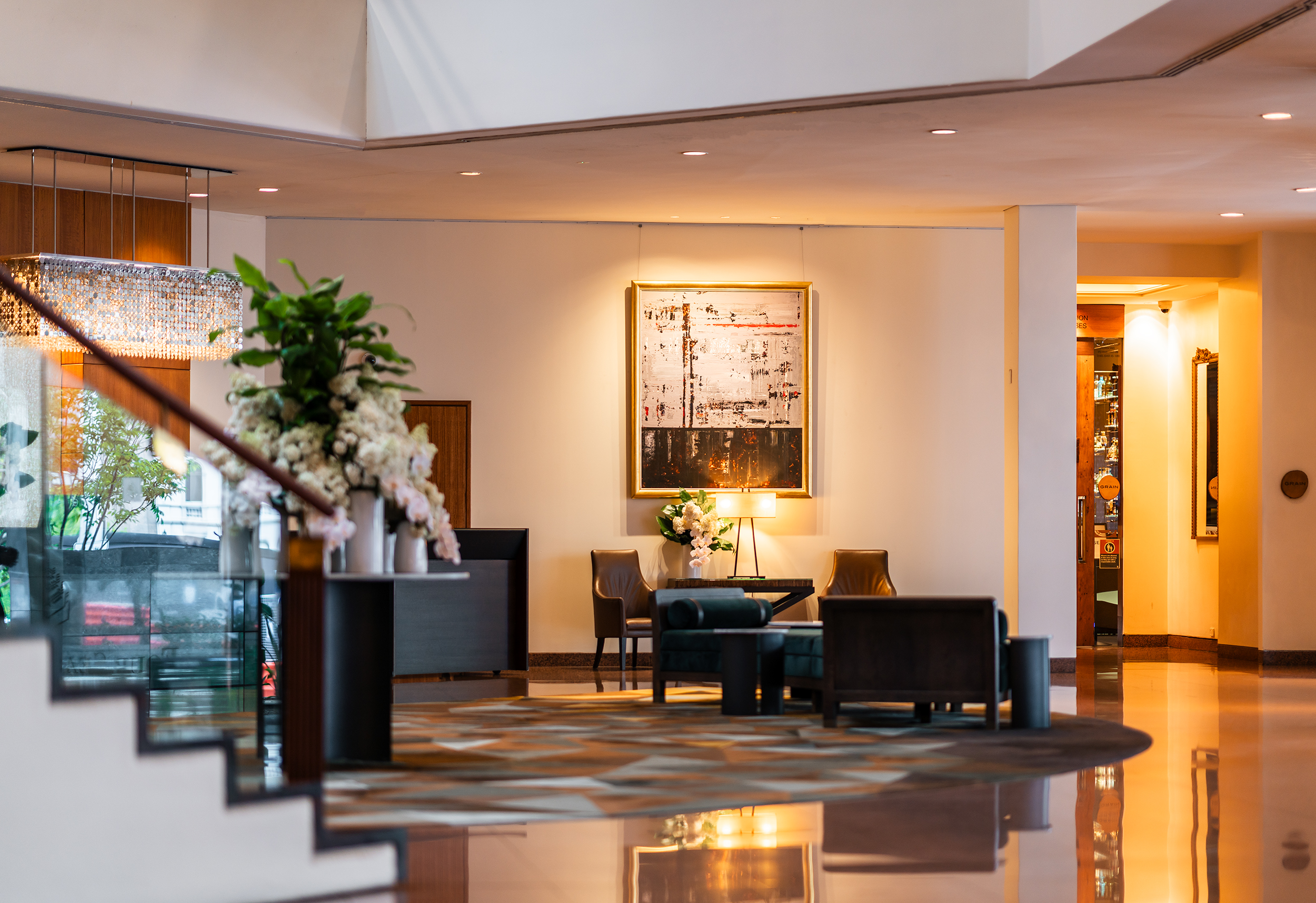 The elegant lobby of the Four Seasons Sydney with a sunken seating area centered on a circular rug, featuring deep green armchairs and a low marble coffee table. A modern abstract painting in a gold frame hangs above a pair of leather side chairs, while a floral arrangement on the reception desk adds a touch of freshness