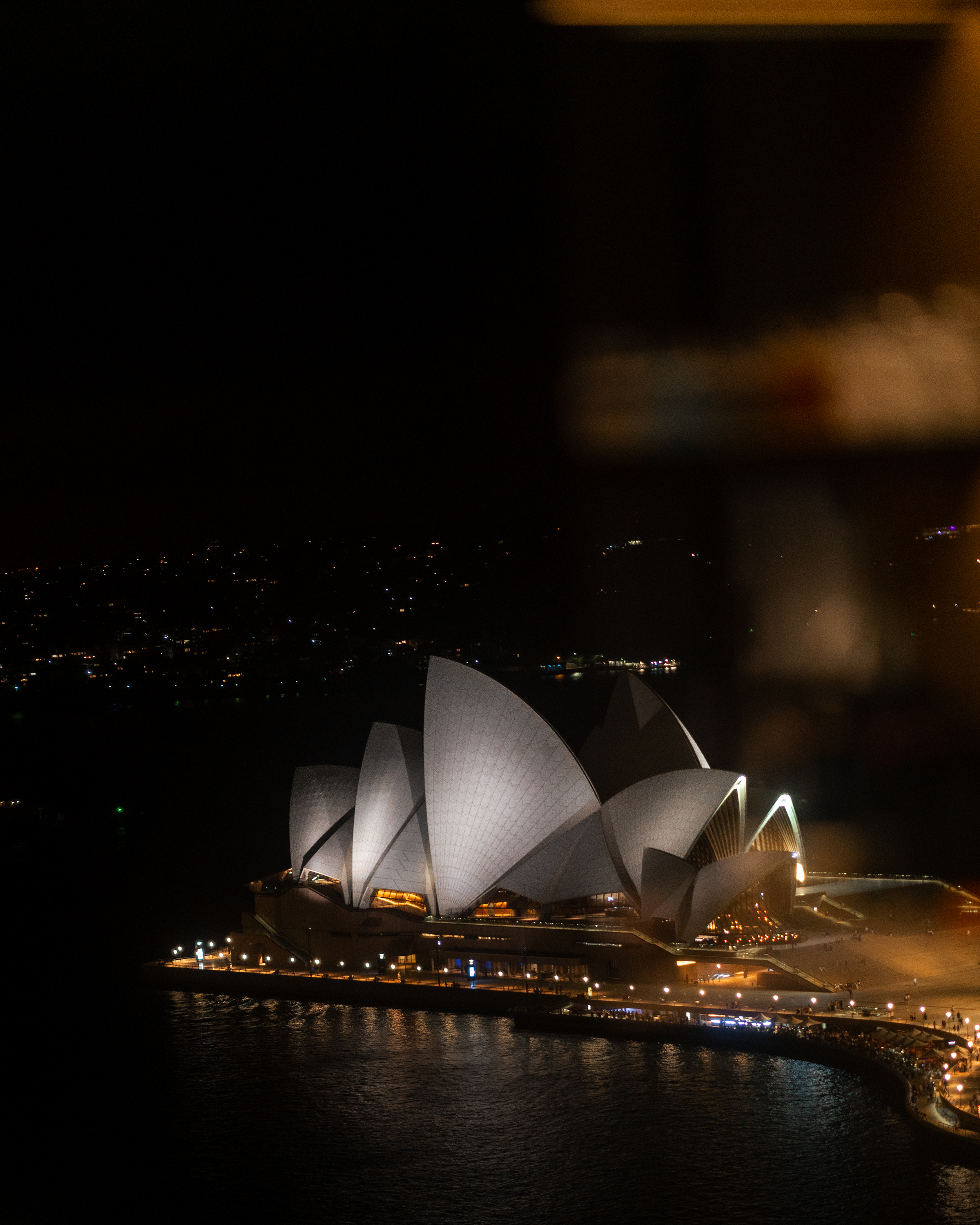 A nighttime view from a high-rise Four Seasons Sydney guest room window capturing the illuminated sails of the Sydney Opera House against a dark harbor backdrop. Reflections of in-room lighting blur softly on the glass, framing the iconic architectural landmark