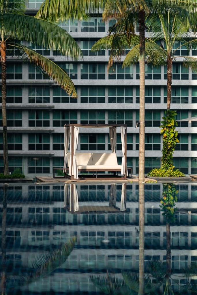 A cream-canopied daybed framed by tall palm trees, mirrored in the still water of the infinity pool against the backdrop of the hotel’s glass-paneled façade in soft morning light