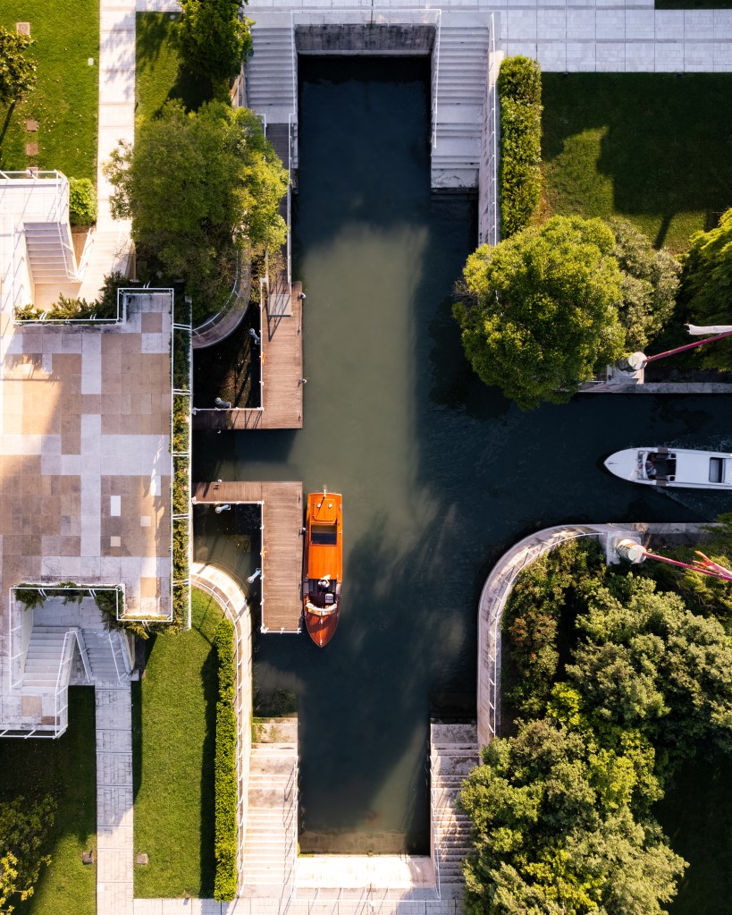Overhead drone perspective of a polished wooden launch tied to a private dock in front of the JW Marriott Venice, lagoon water lapping at stone terraces