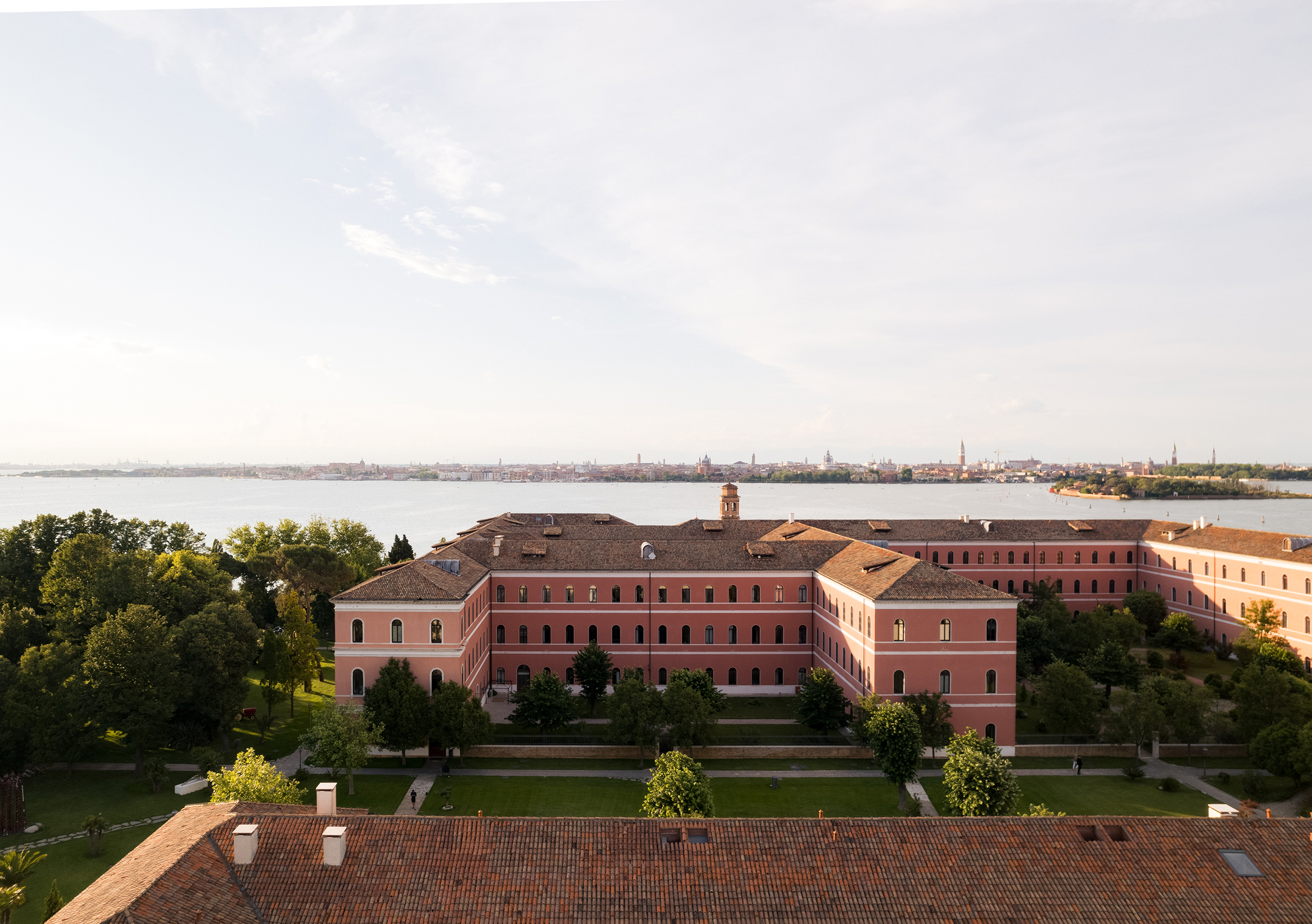 A wide aerial panorama of Venice at midday: the terracotta rooftops and historic buildings of the island city stretch across the horizon, framed by the calm blue expanse of the lagoon