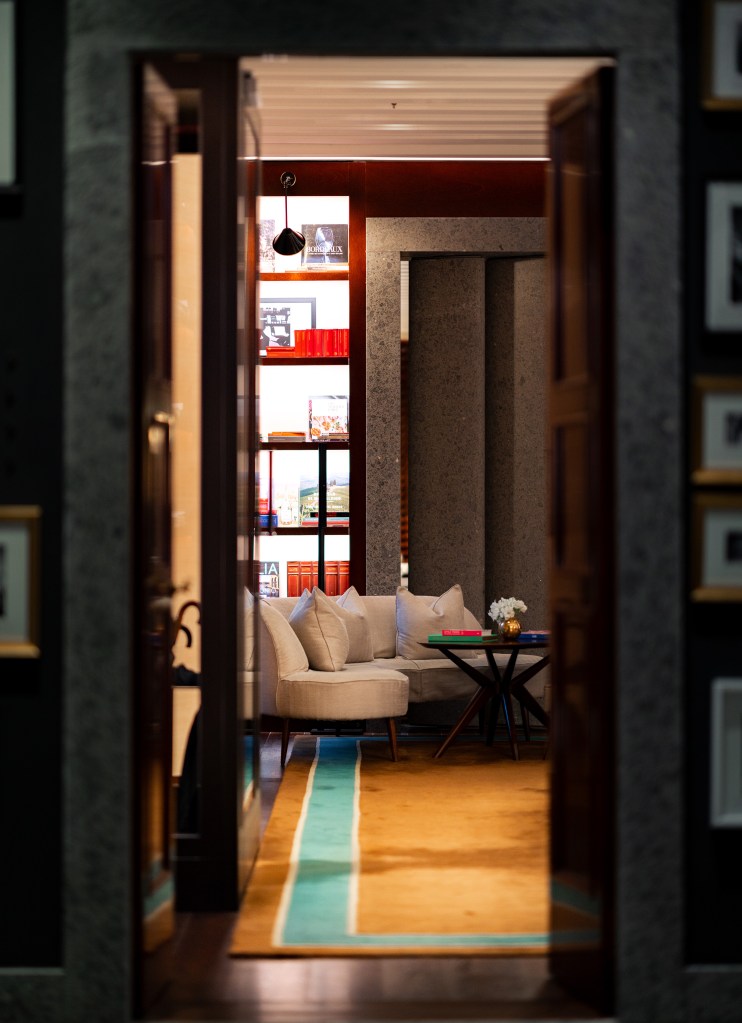 View through a dark wood doorway into an intimate hotel lounge: curved beige sofa around a small round table with books and flowers, backlit floor-to-ceiling shelving filled with books and decorative objects