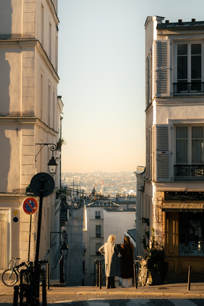 Golden hour street scene in Montmartre, Paris, with two people talking at the top of stairs and hazy city skyline in the distance
