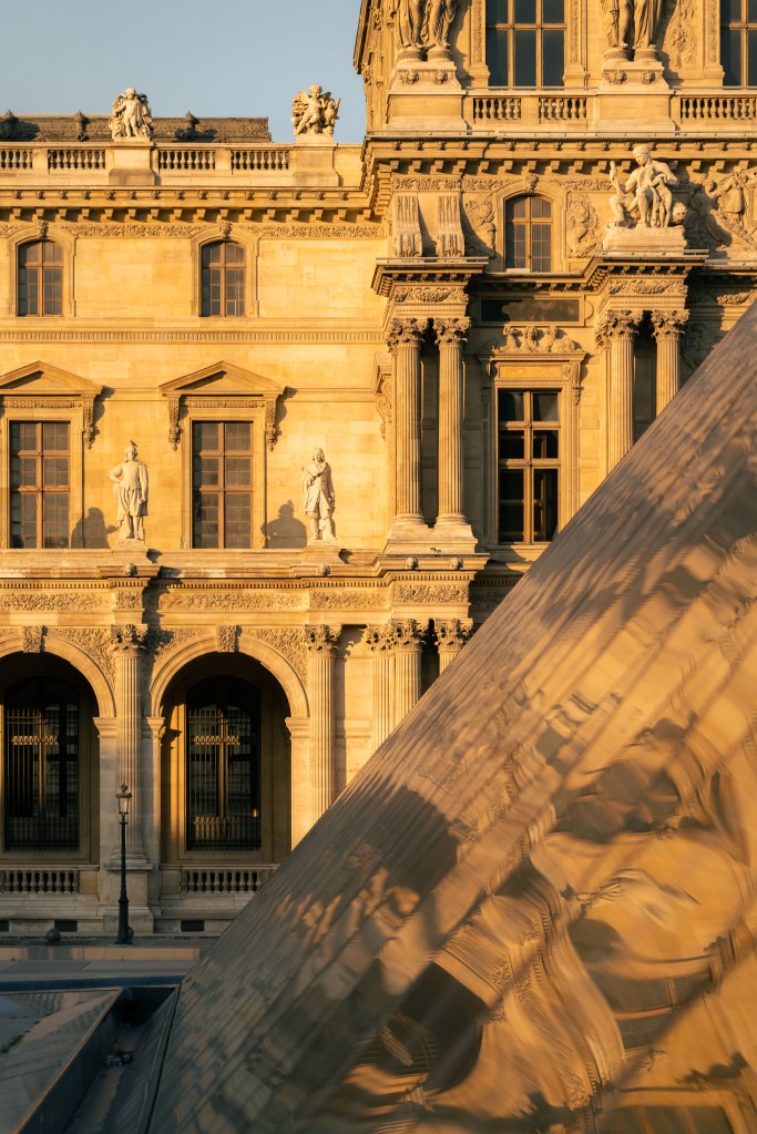 Louvre Museum façade in Paris with ornate stone arches and statues, seen behind the glass pyramid catching soft golden sunset reflections