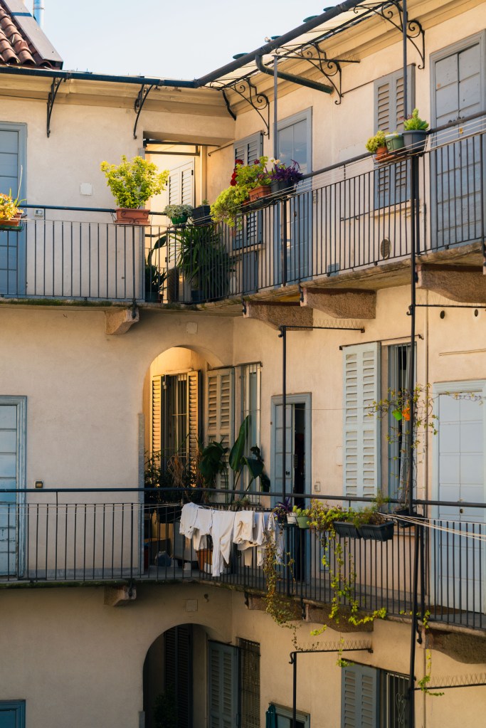 Sunlit Milan apartment courtyard with pale walls, blue shutters, potted plants on iron balconies and white laundry drying on the railing