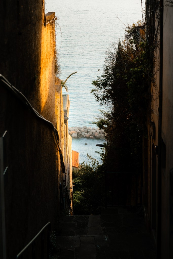 Narrow shaded alley in Marseille leading downhill toward the Mediterranean, with golden light on one wall and a glimpse of rocks and sea at the end