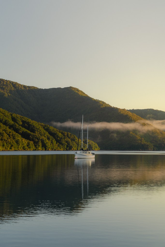 Sailboat anchored on still water near Picton, New Zealand, with forested hills, a low band of mist and soft golden morning light
