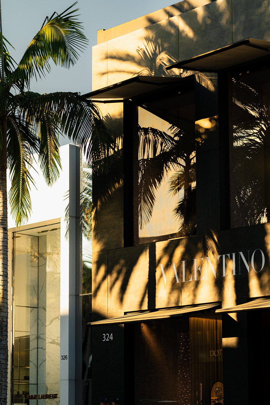 Golden hour sunlight casting palm tree shadows across the black Valentino storefront on Rodeo Drive, with the neighboring Saint Laurent boutique visible in the background.
