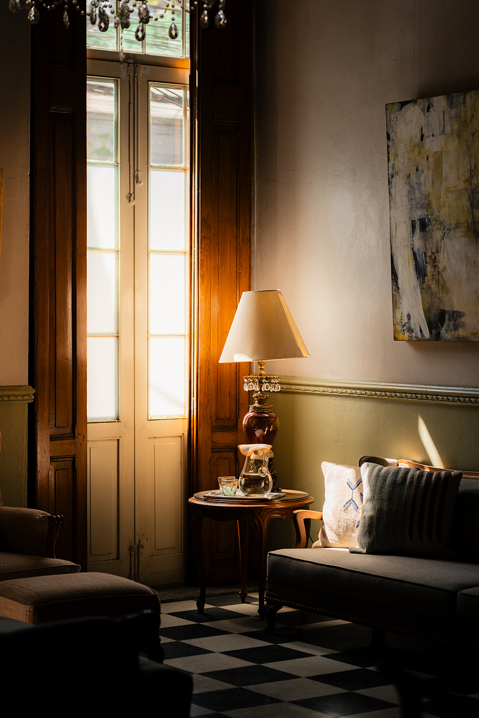 Warm afternoon light streaming through tall wooden French doors onto a vintage lamp, side table with a carafe of water, and a grey sofa with cushions, in a cozy sitting room with black-and-white checkered floor at El Patio 77 in Mexico City