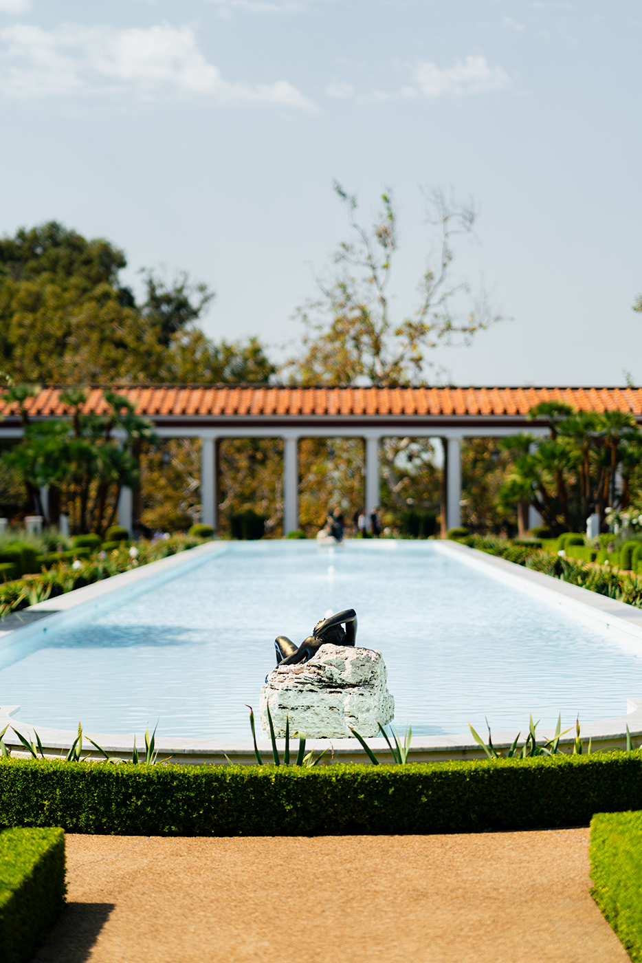 Long reflecting pool at The Getty Villa near Los Angeles, with a small sculpture of reclining legs on a rock in the foreground and a colonnade with terracotta roof and manicured gardens in the background.