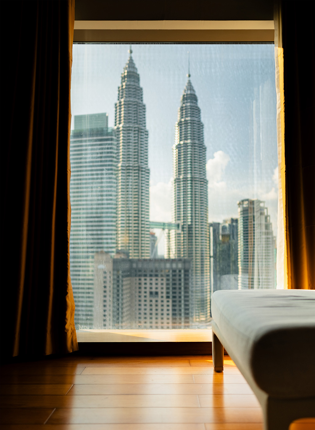 View from a Grand Hyatt Kuala Lumpur room with the Petronas Towers framed in the window, warm light on the wooden floor and a soft daybed in the foreground.