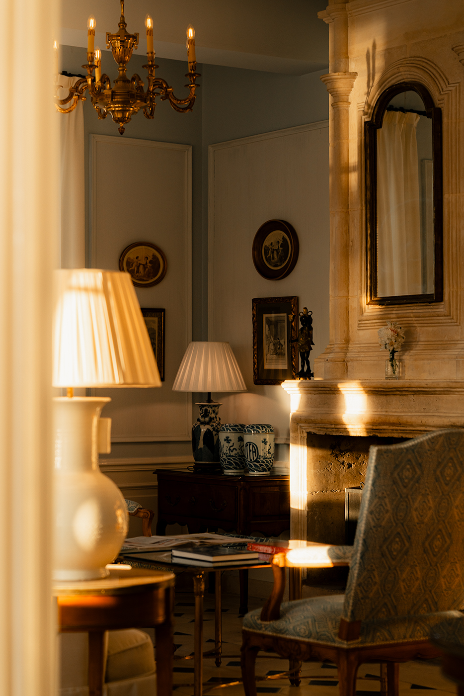 ntimate interior of Hôtel Le Toiras suite: warm lamp light spills onto an antique marble fireplace, gilded frames, and a richly upholstered armchair.