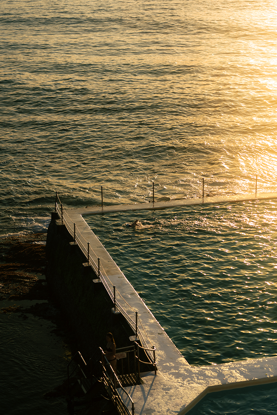 Golden hour view of the Bondi Icebergs ocean pool with one swimmer in the lane and a woman standing on the pool stairs, framed by shimmering sunlit sea