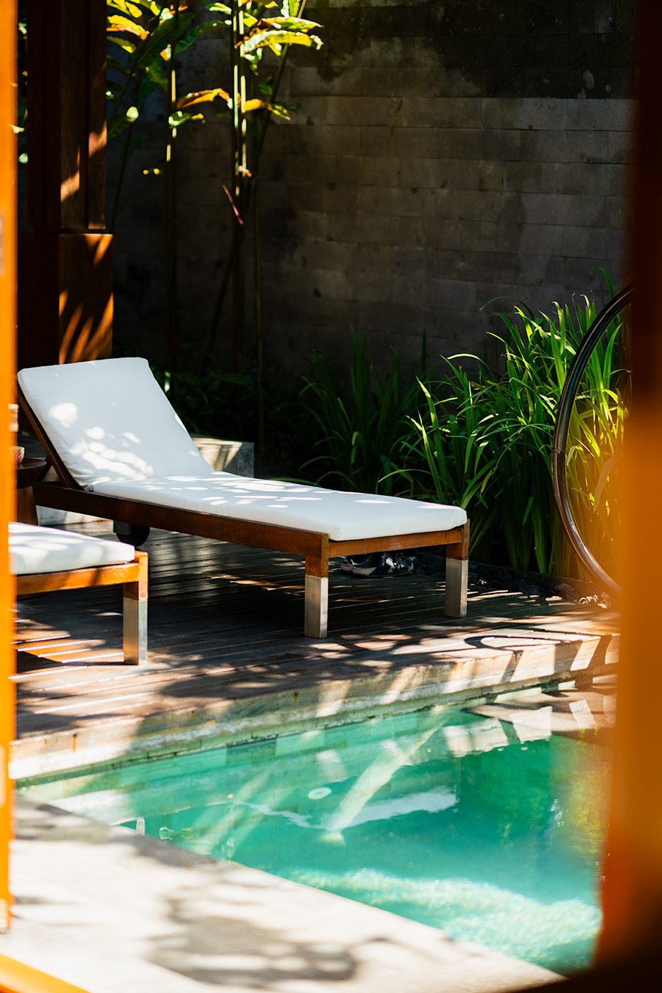 Sunlit private villa terrace at Indigo Bali with a white cushioned lounger by a turquoise plunge pool, surrounded by lush green plants and soft shadow patterns on the wooden deck.