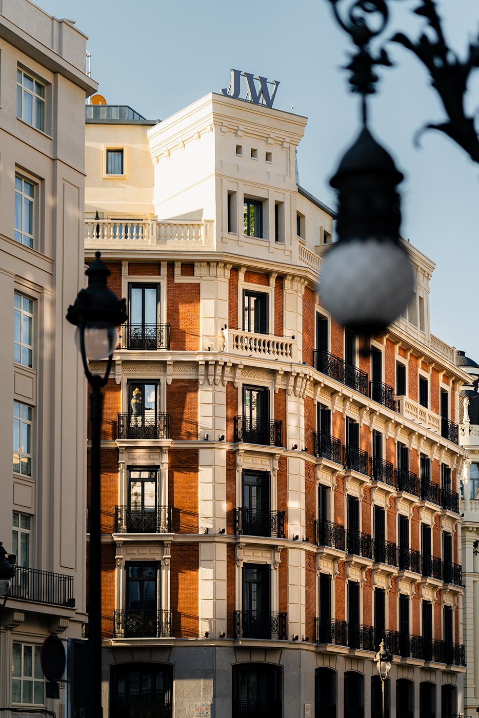 Late-day light illuminating the hotel’s elegant brick and stone corner building topped with a “JW” rooftop sign, framed in the foreground by out-of-focus wrought-iron street lamps