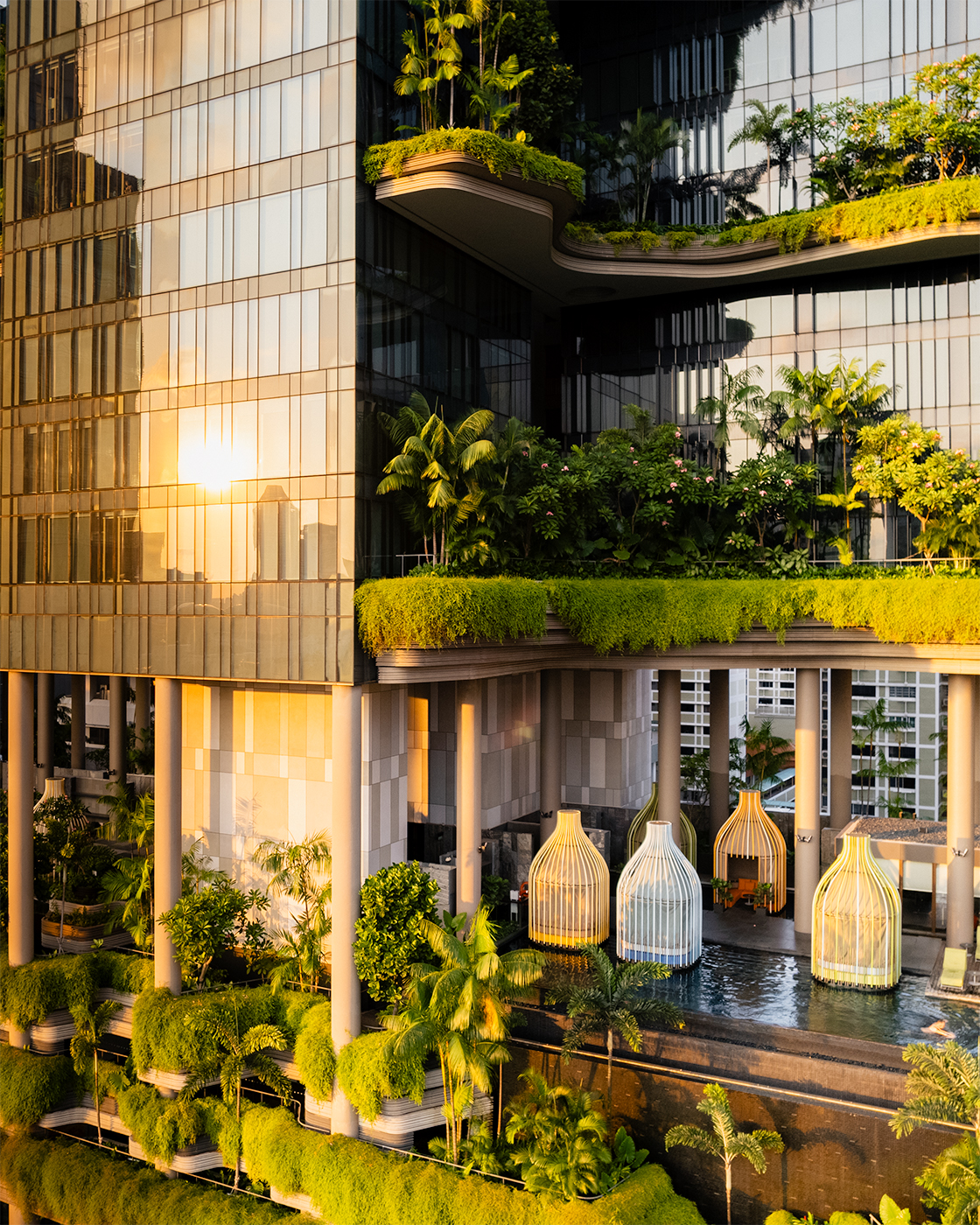 Aerial sunrise view of PARKROYAL COLLECTION Pickering in Singapore, with tiered sky gardens and glass façade glowing in warm golden hour light above the poolside birdcage cabanas