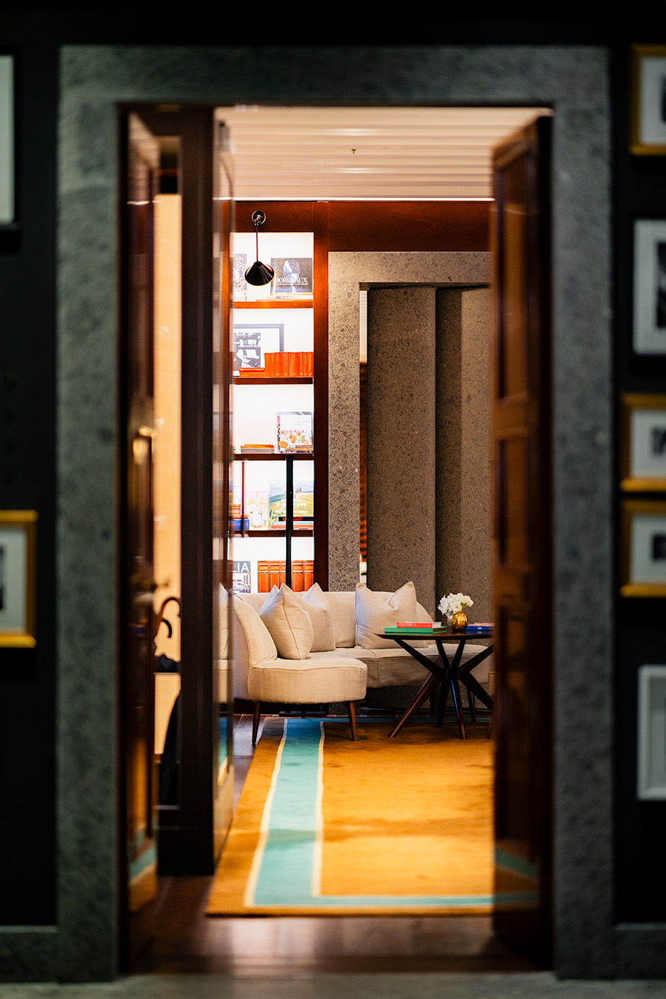 View through a dark wood doorway into a quiet reading nook, where a curved cream sofa beneath built-in bookshelves is lit by hidden LED strips, accented by a small round marble-topped table holding fresh flowers