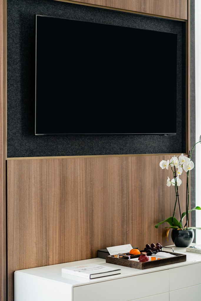 Wall-mounted TV above a white cabinet with an orchid and a welcome tray of fruit and desserts in a modern hotel room