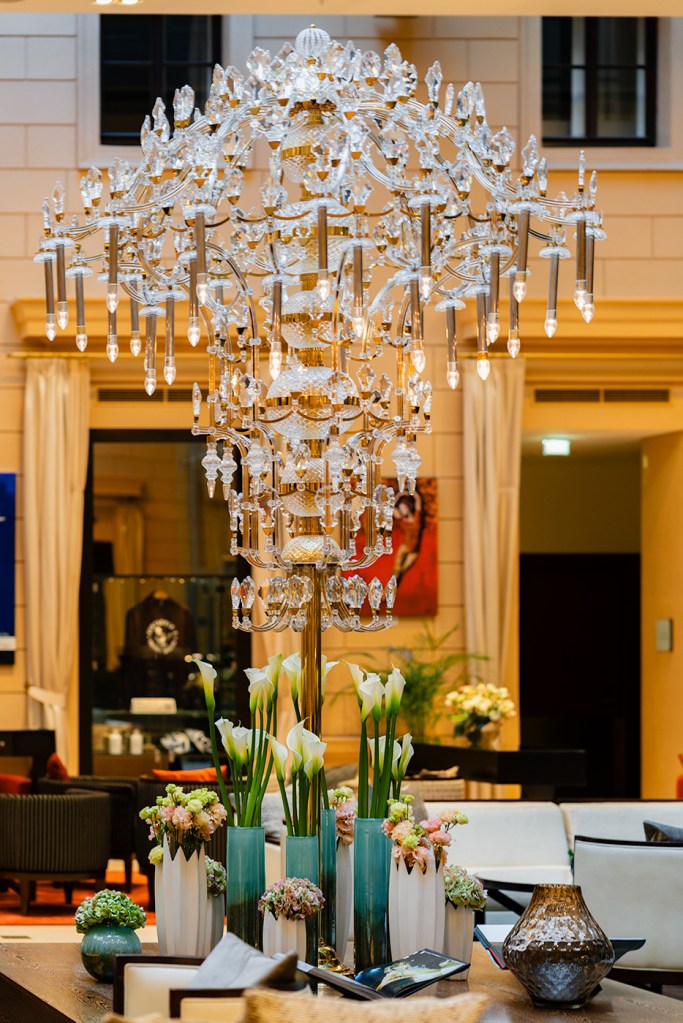 Dramatic crystal chandelier centerpiece rising from a lobby table surrounded by tall vases of calla lilies and pastel floral arrangements