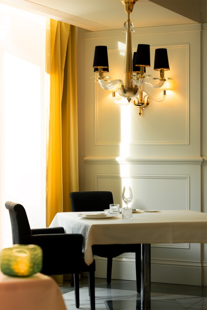 Sunlit restaurant corner with a set table beneath a brass chandelier, black chairs, white wall molding, and yellow curtains.