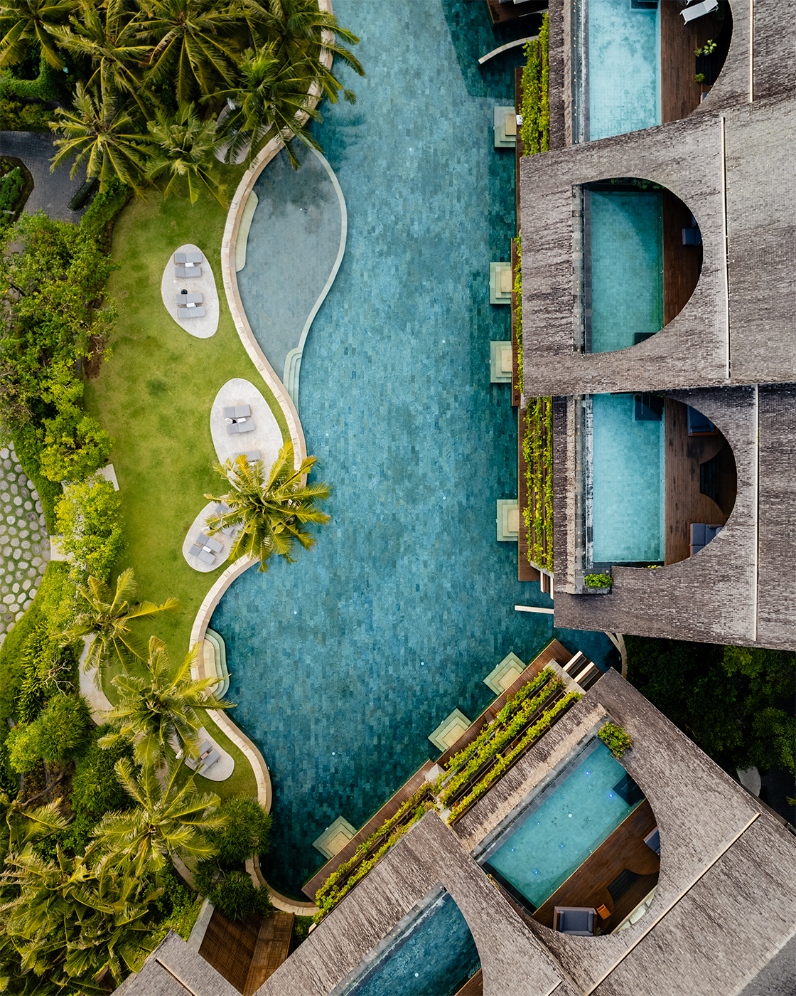 Aerial drone view of the main lagoon pool and private plunge pools at COMO Uma Canggu in Bali, framed by palm trees, green lawns and modern rooftop suites