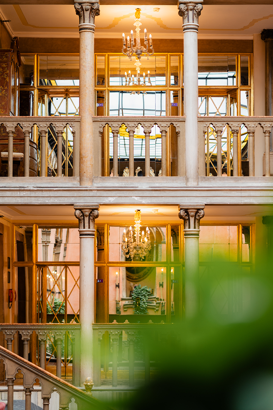 Interior atrium view at Hotel Danieli in Venice, showing columns, balustrades, chandeliers, and reflective golden window frames, with soft greenery in the foreground