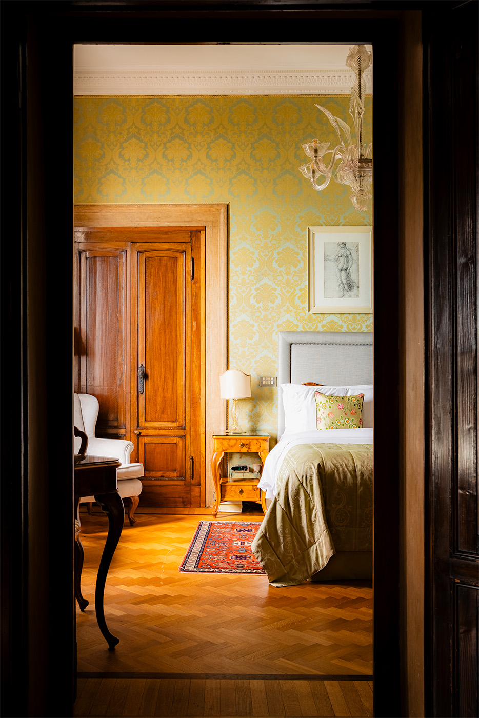Doorway view into a classic Hotel Danieli bedroom in Venice, featuring patterned wallpaper, wooden doors, a made bed with a green throw, and a small chandelier.