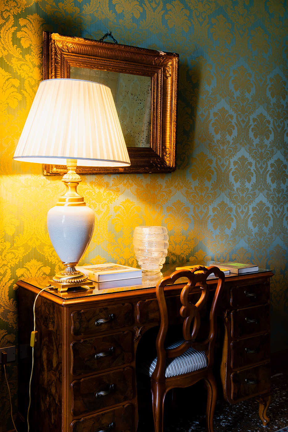Antique writing desk and carved chair at Hotel Danieli in Venice, lit by a white table lamp beside a gilded mirror on damask wallpaper.
