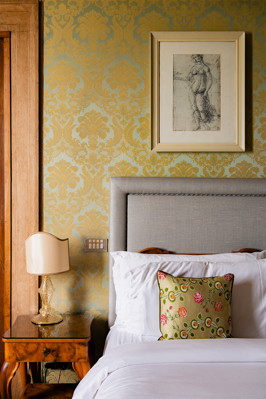 Bedside detail in a Hotel Danieli room in Venice with damask wallpaper, framed artwork above the headboard, a glass-base bedside lamp, and a floral accent cushion