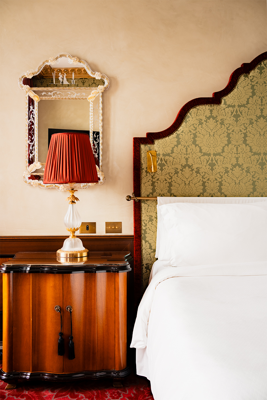 Elegant bedside vignette at Hotel Danieli in Venice with an ornate mirror, red pleated lamp, polished wood cabinet, and crisp white bedding