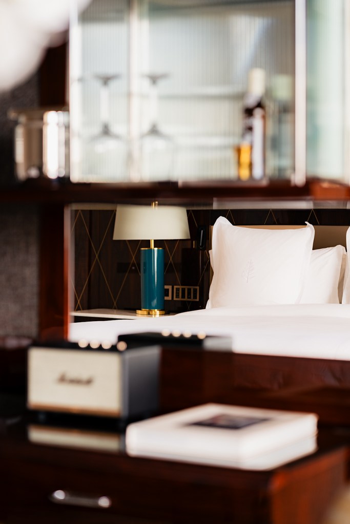 Four Seasons Lisbon guest room detail seen through a minibar shelf, with crisp white bed linens and a teal bedside lamp, books and a speaker softly blurred in the foreground.