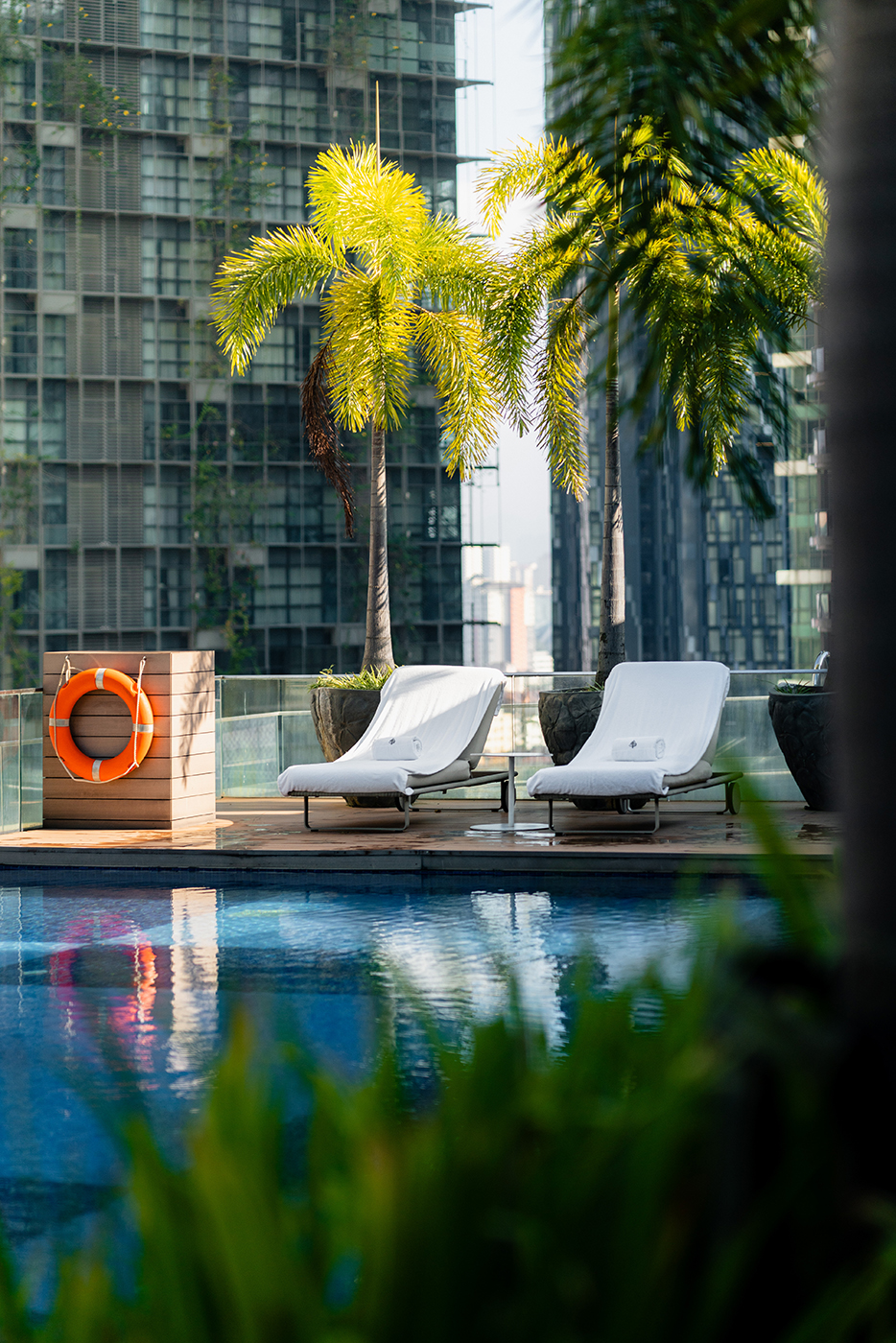 Four Seasons Kuala Lumpur pool deck with two white loungers under palm trees, reflected in the water, with high-rises behind