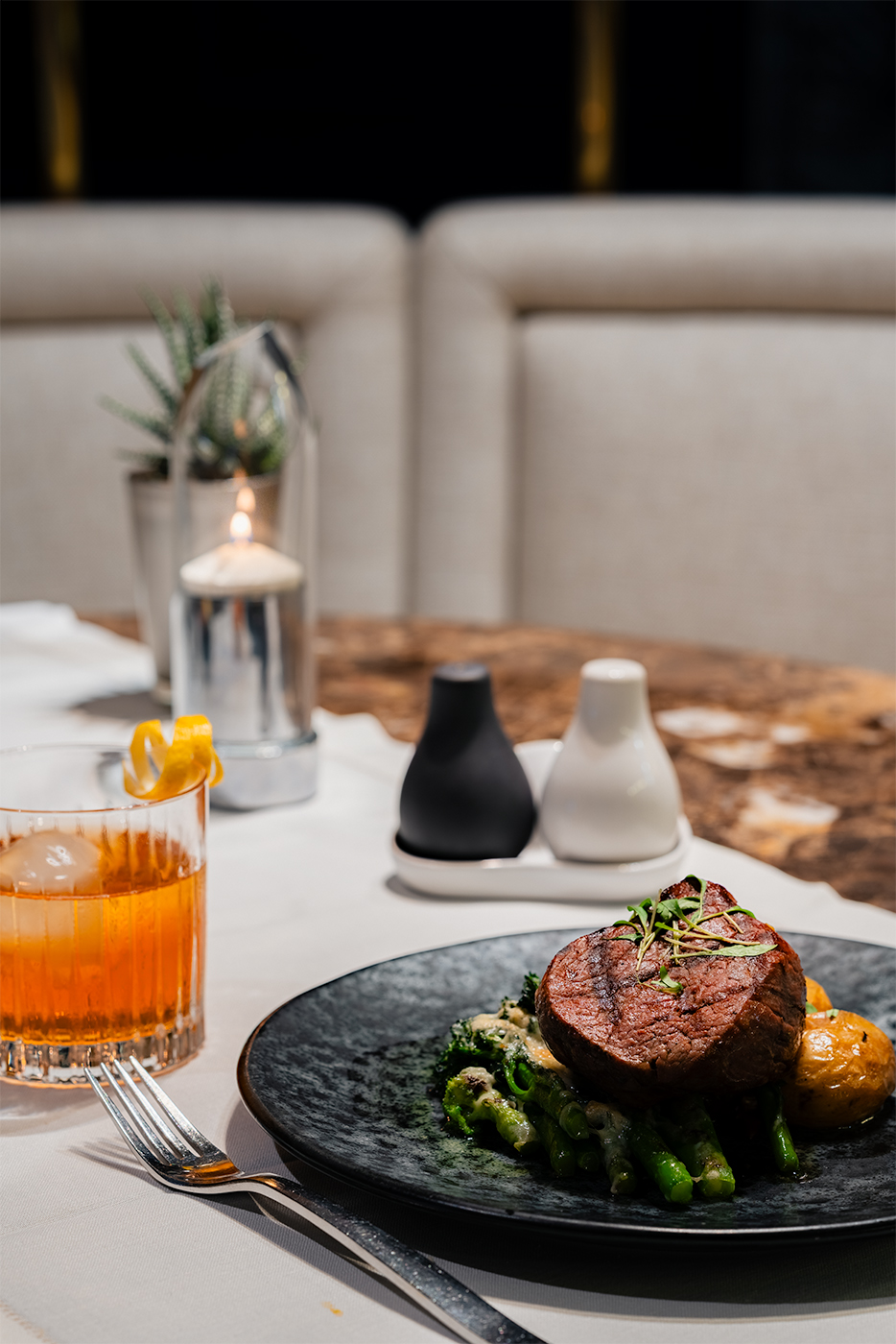 Plated steak with asparagus on a black plate beside an amber cocktail and candlelit table setting, InterContinental Lisbon