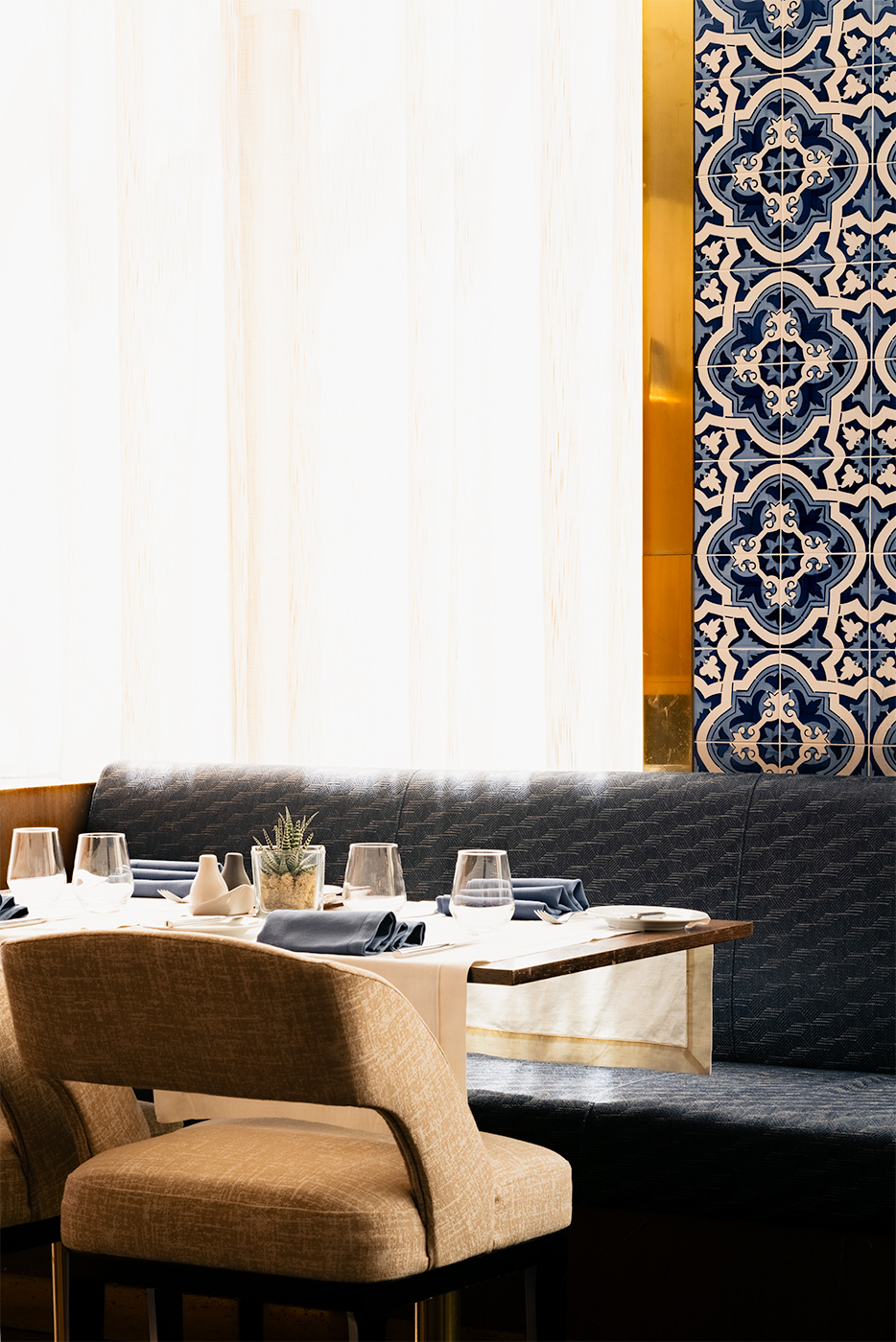 Restaurant banquette setting in Lisbon with blue patterned tiles, soft daylight through sheer curtains, and a neatly set table with glassware