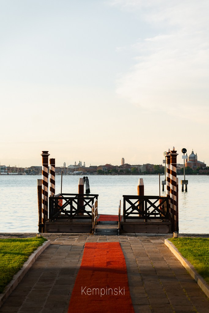 Red-carpeted pier with striped mooring poles leading to the lagoon, Venice skyline in the distance at sunset