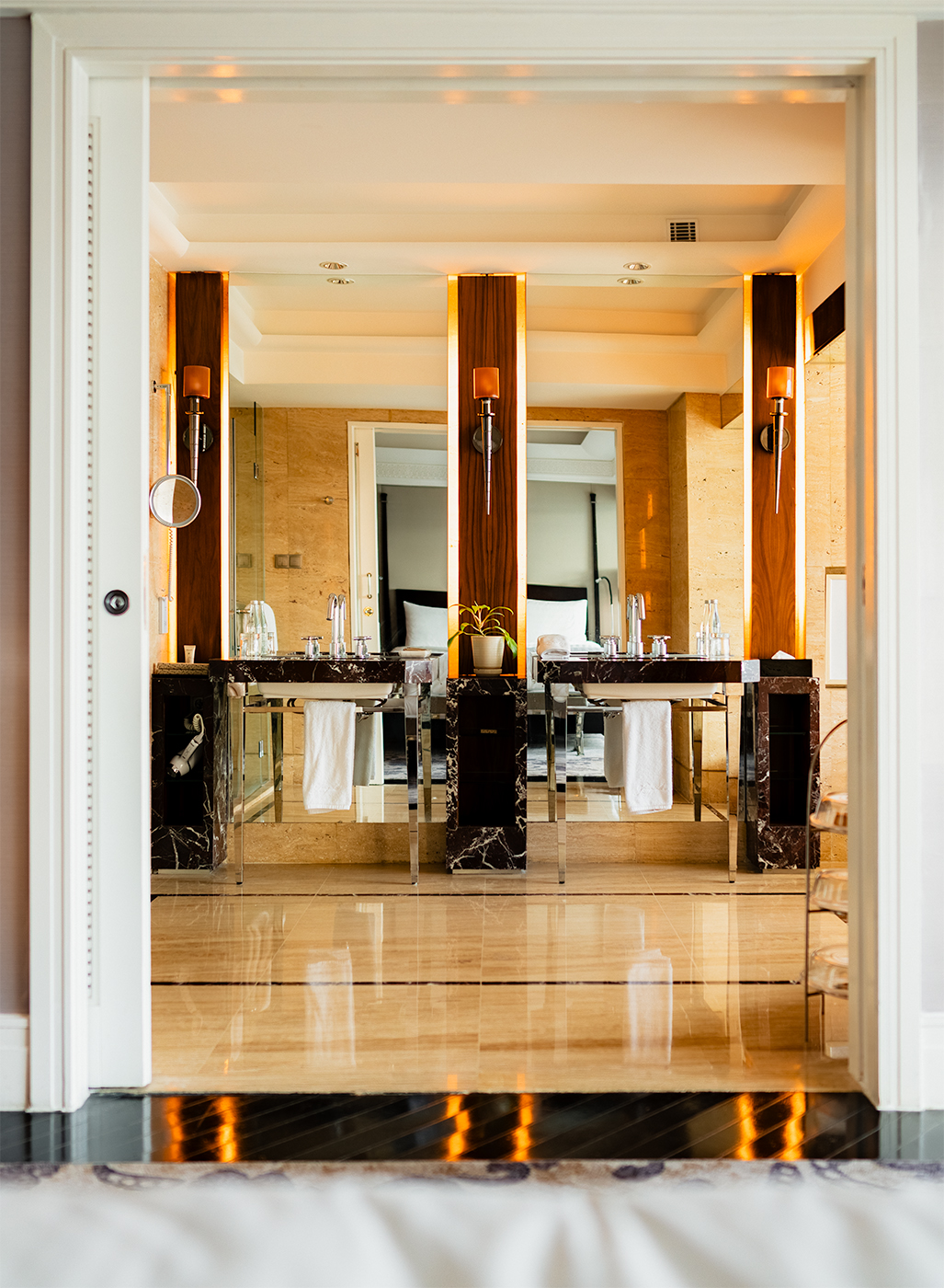 Symmetrical double-sink bathroom vanity with backlit mirrors and warm wood panels, viewed through a doorway