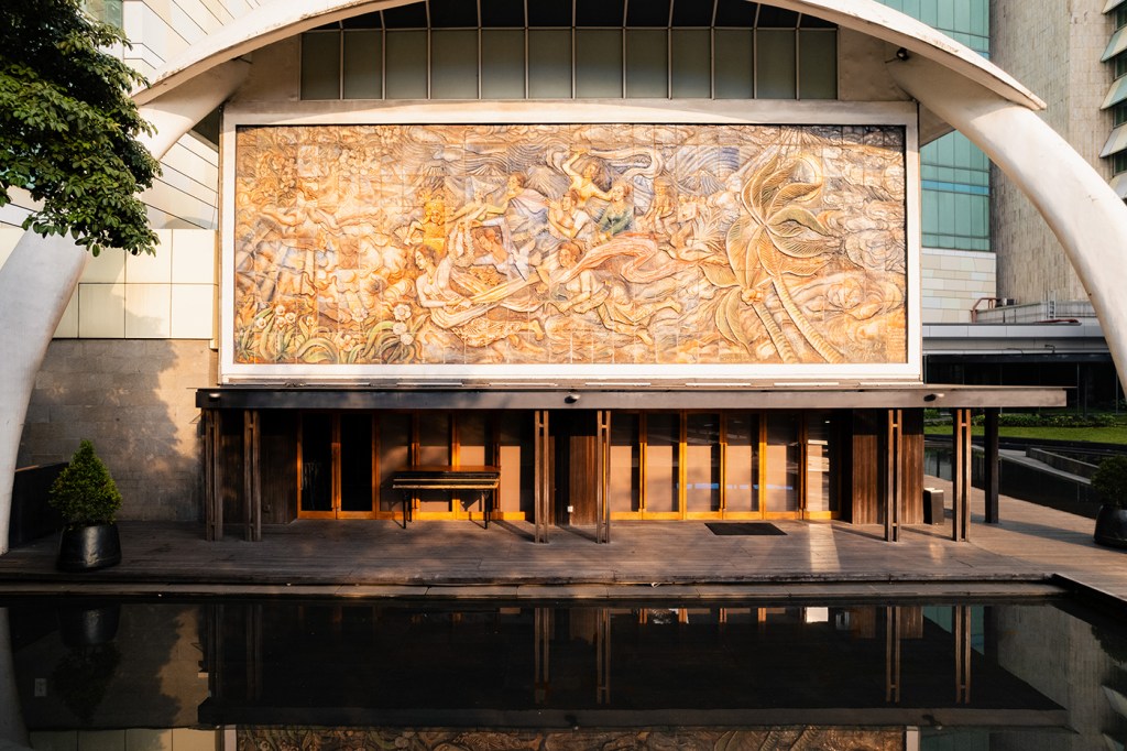 Hotel entrance beneath a curved arch with a large mural facade and a reflecting pool in warm evening light