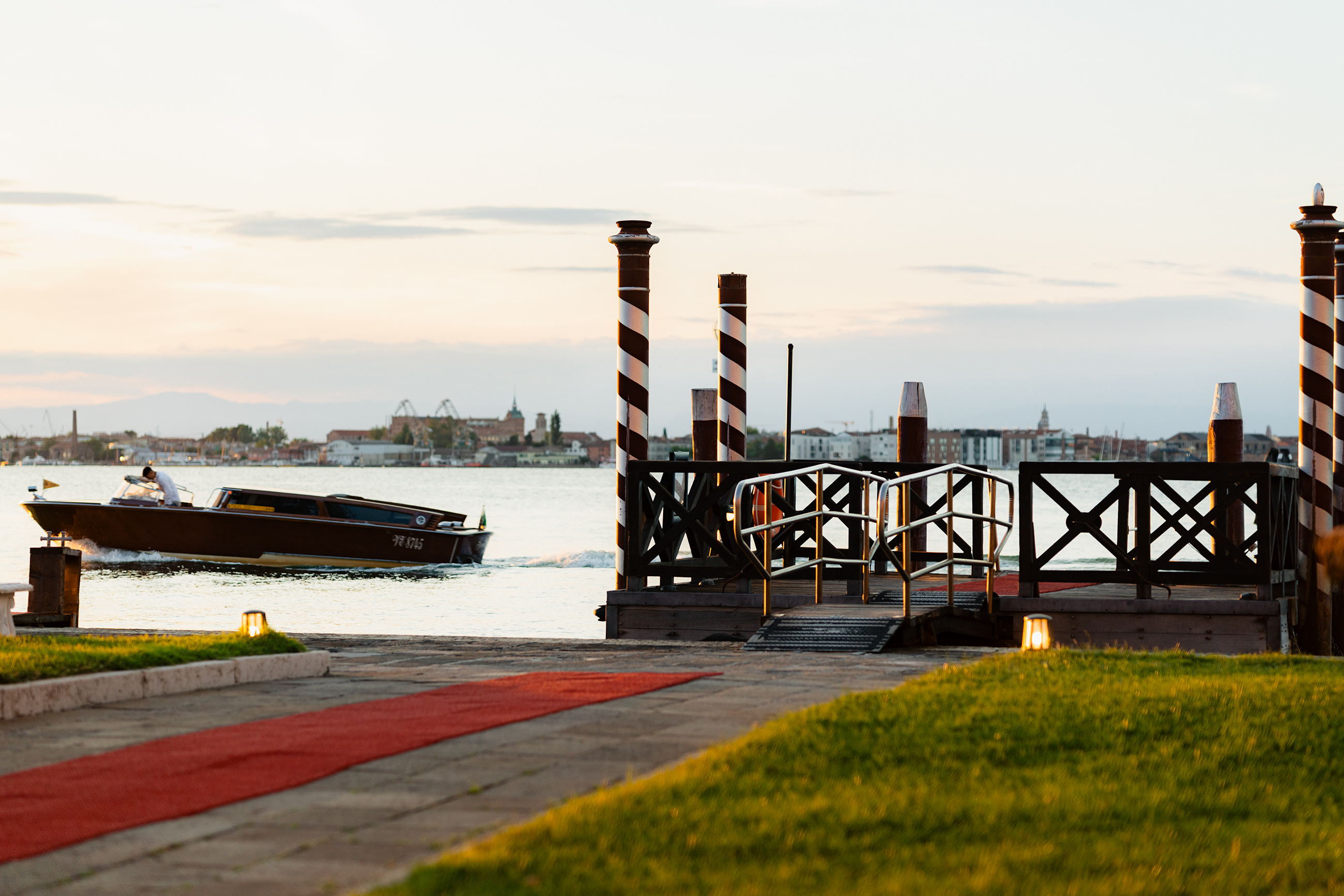 Lagoon landing stage with striped poles and red carpet, with a water taxi passing in the background at dusk