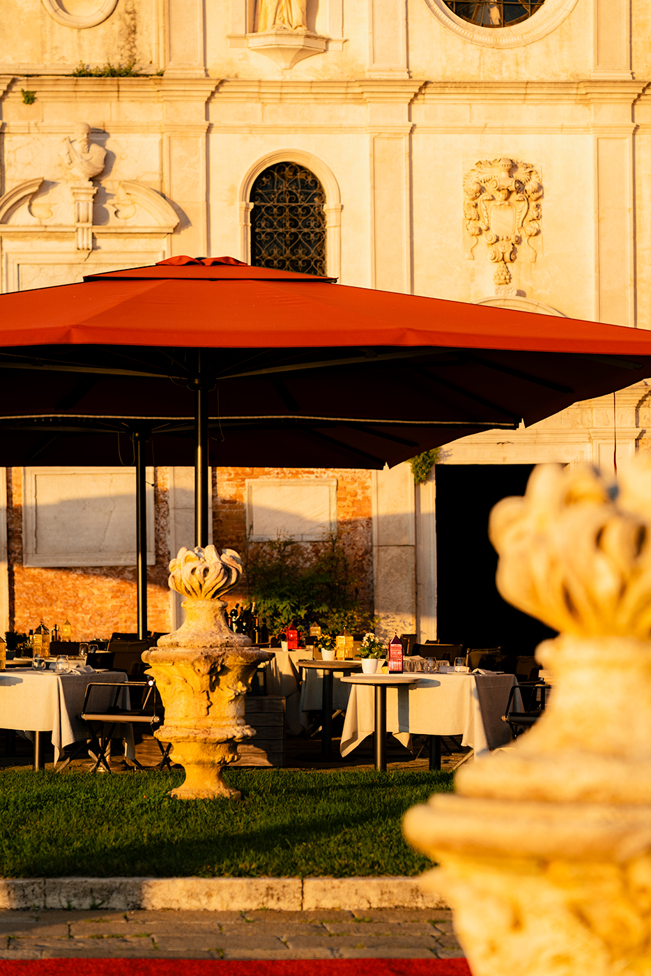 Outdoor dining terrace under a large red umbrella with set tables against a historic facade in golden light.