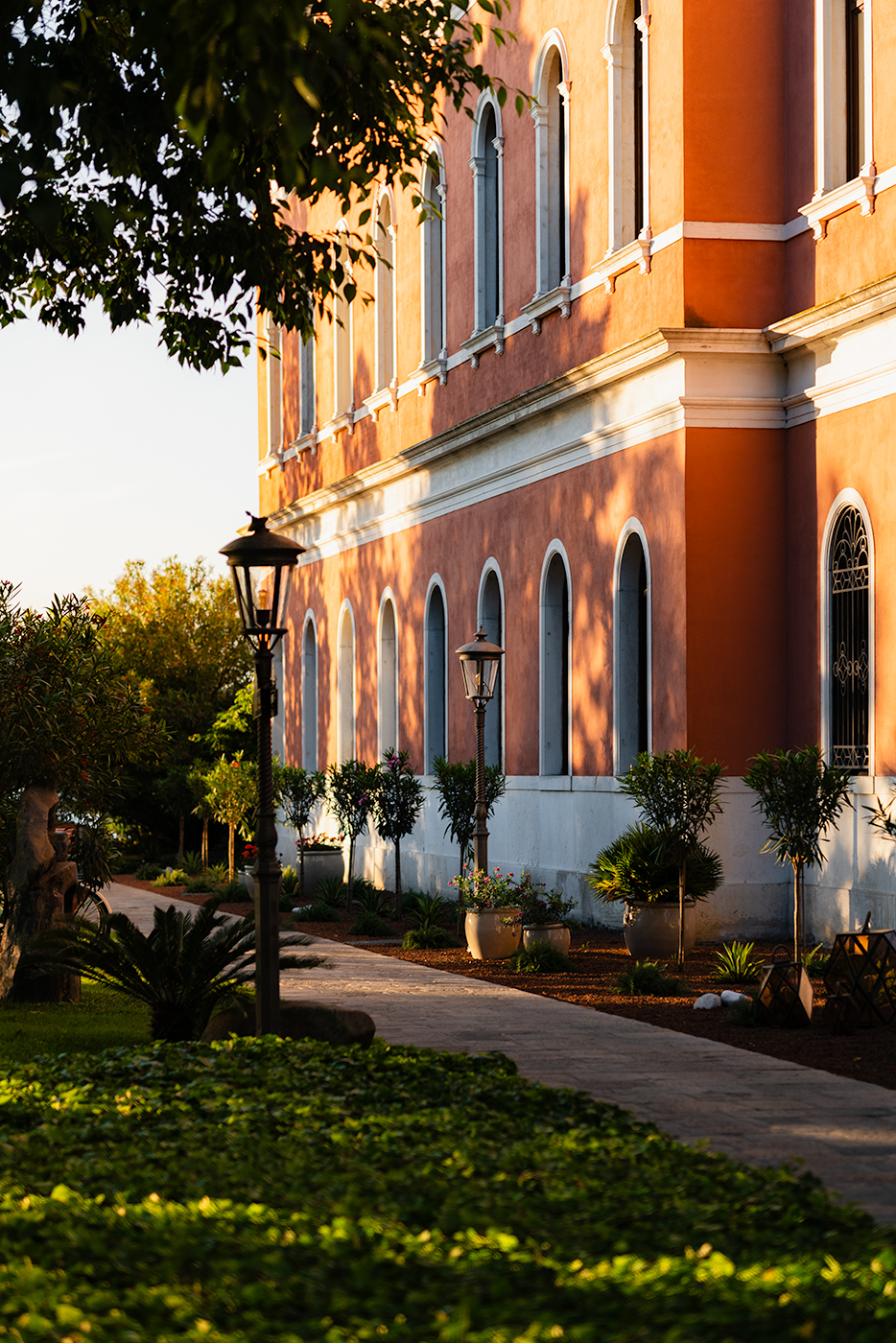 Garden path alongside a terracotta building with arched windows and lampposts in warm evening shadows