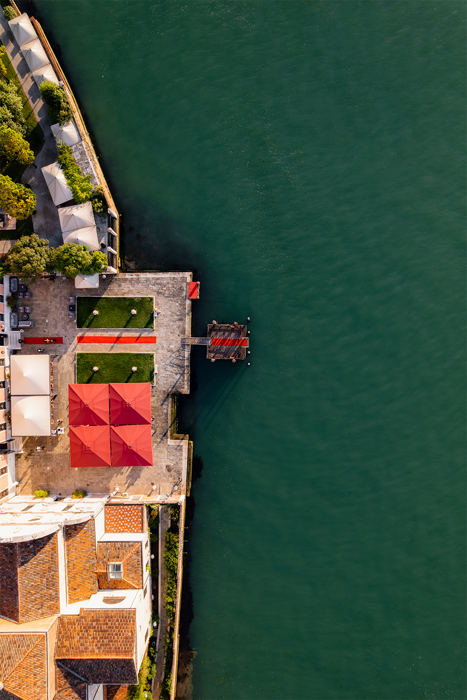 Aerial view of a waterfront terrace with red umbrellas and a pier extending into the green lagoon.