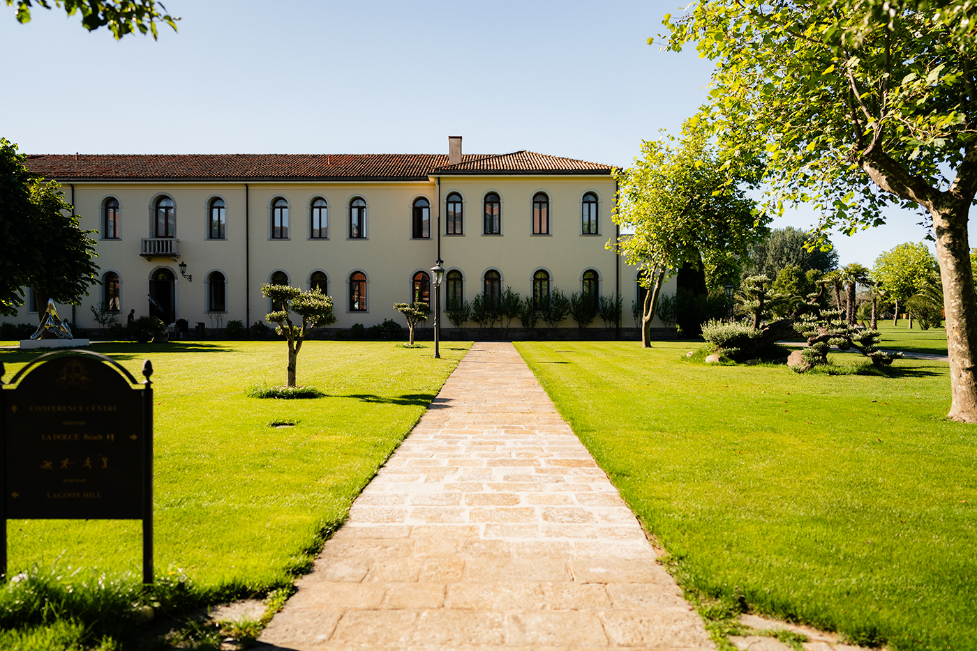 Long hotel facade with arched windows and a central stone walkway across a manicured lawn